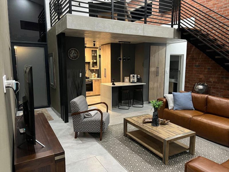 Modern open-plan living area featuring a double-volume ceiling, exposed brickwork, a mezzanine level, and a contemporary kitchen with a central island.