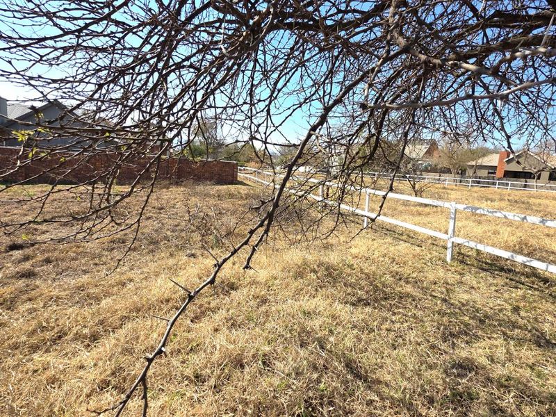 A vacant residential plot featuring dry grass, a white picket fence, and a brick boundary wall.