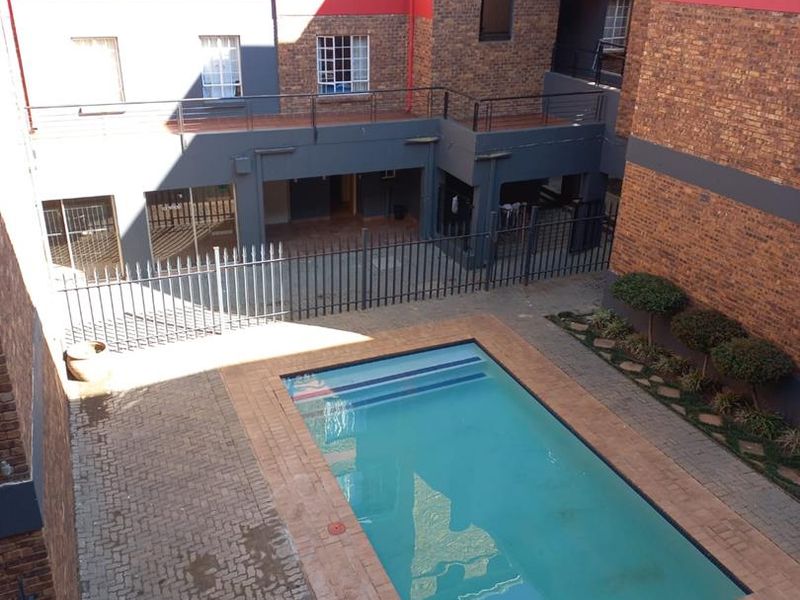 An elevated view of a communal swimming pool area featuring brick paving, security fencing, and surrounding apartment building facades.