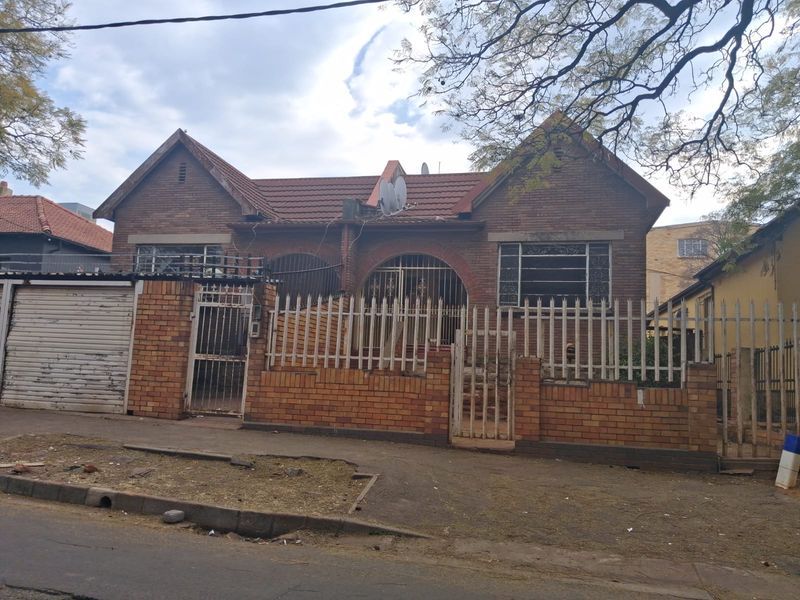 Front elevation of a single-story brick house featuring a tiled roof, security fencing, and an integrated garage, showing signs of age and wear.