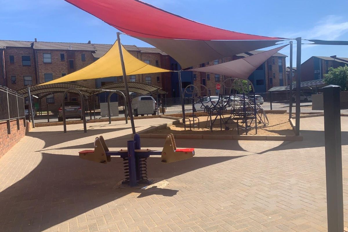 Communal outdoor play area featuring paved ground, sand pit, playground equipment, and overhead shade sails within an apartment complex.