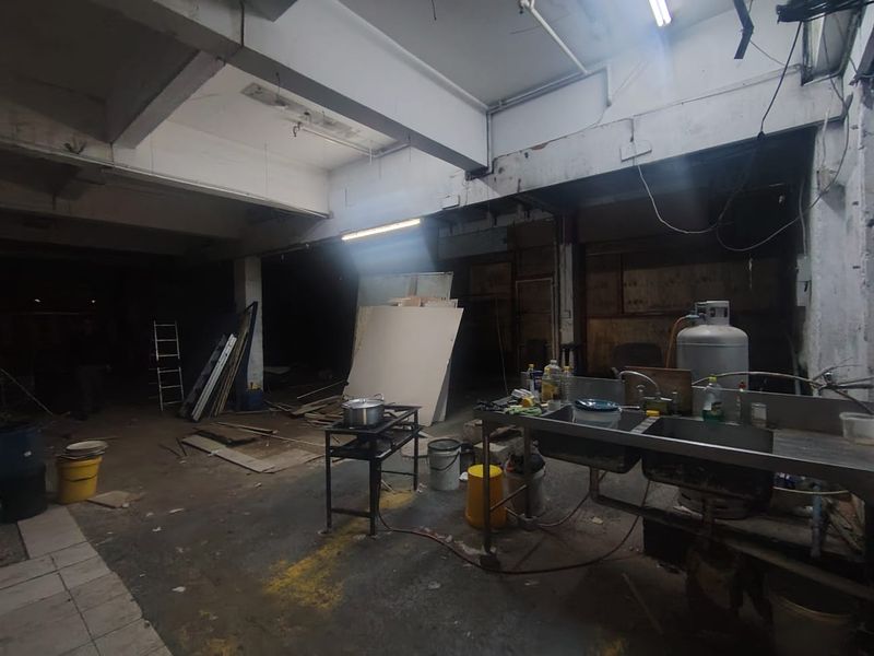 An industrial utility space featuring exposed concrete beams, basic lighting, and a stainless steel sink unit, currently in a shell-like condition.