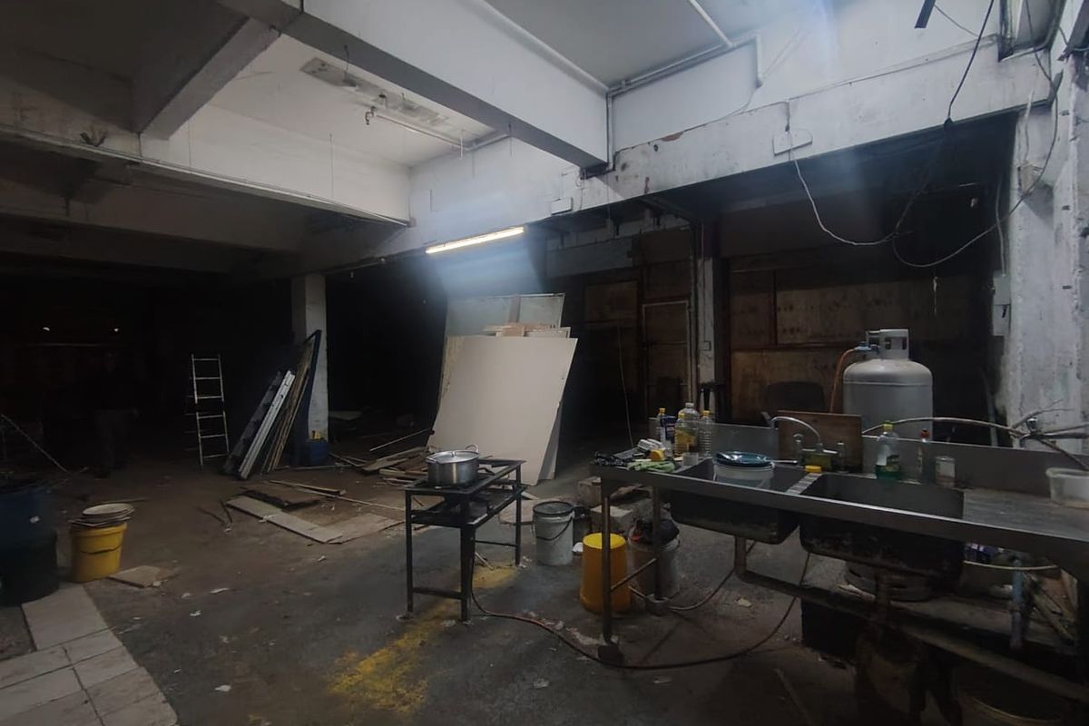 An industrial utility space featuring exposed concrete beams, basic lighting, and a stainless steel sink unit, currently in a shell-like condition.