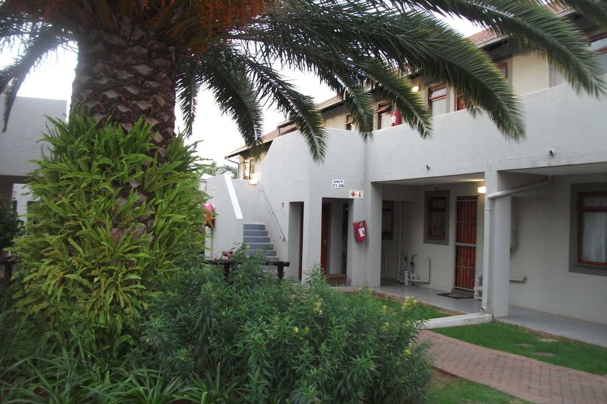 Exterior view of an apartment complex featuring a paved walkway, landscaped garden with a large palm tree, and access stairs to upper units.