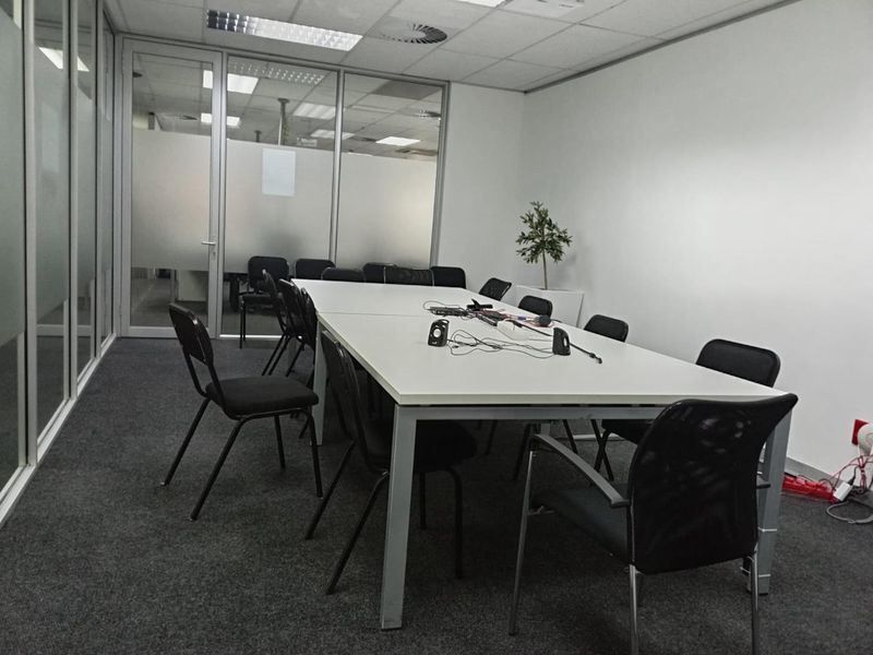 A functional office meeting room featuring glass partition walls, carpeted flooring, and a suspended ceiling with recessed lighting.