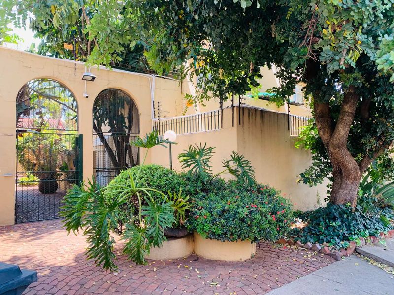 Front entrance area featuring a brick paved driveway, arched metal gate, and perimeter wall with integrated security fencing and established garden planters.