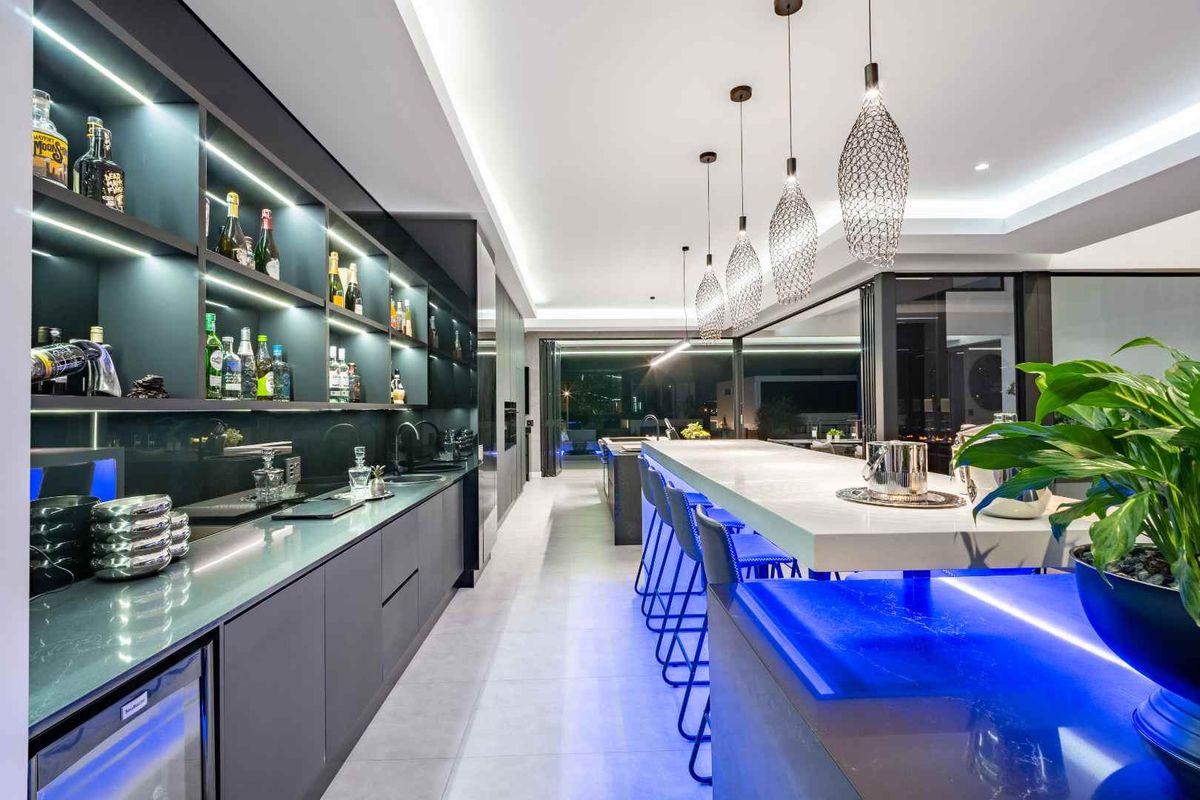 Modern, high-end kitchen featuring sleek dark cabinetry, stone countertops, integrated LED lighting, and a spacious breakfast bar with contemporary pendant fixtures.