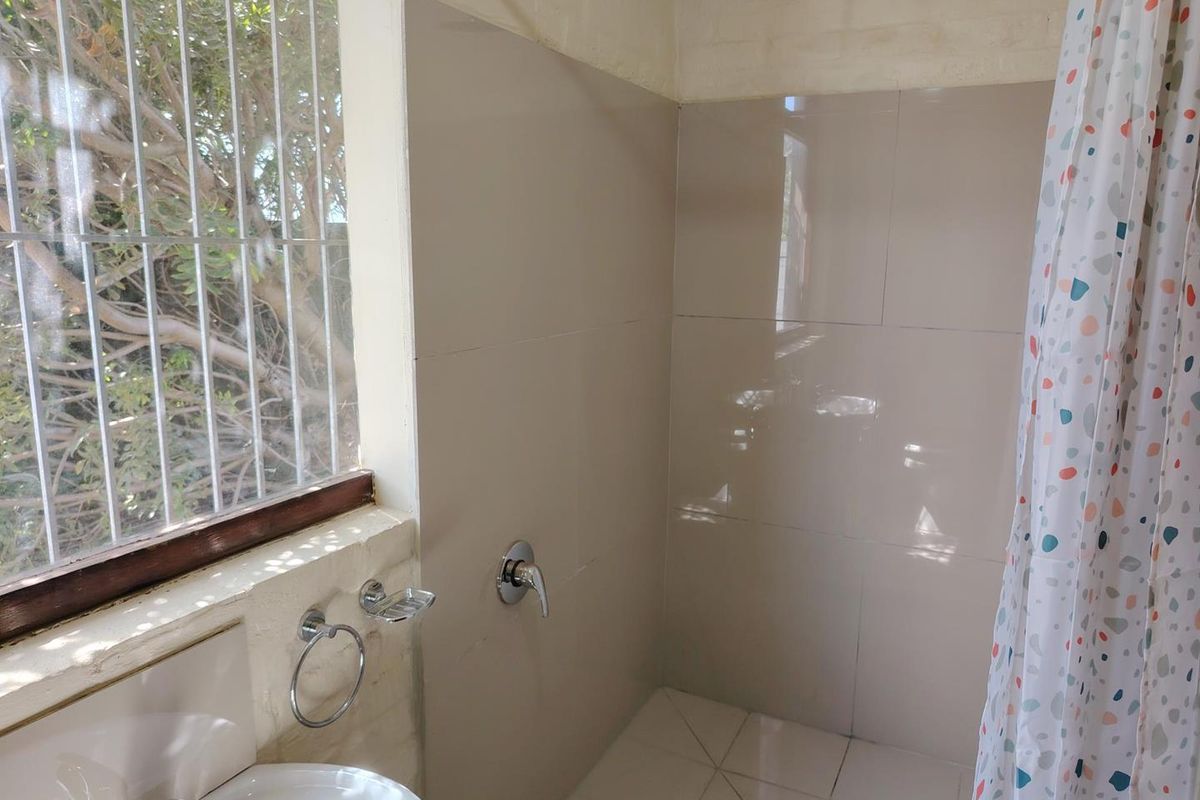 A functional bathroom featuring a shower with wall-mounted mixer, basin, and natural light through a window with security bars.