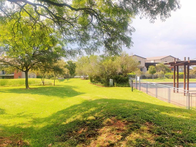 A well-maintained communal garden area featuring expansive green lawns, mature trees, and a fenced pool area with a pergola.