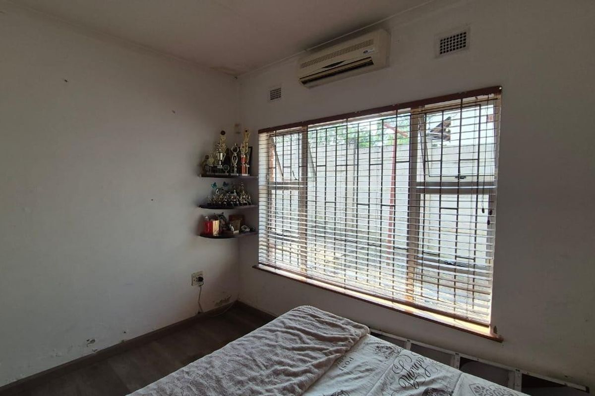 A bedroom featuring laminate flooring, a wall-mounted air conditioning unit, and windows fitted with security bars and blinds.