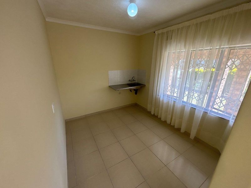 A compact, empty kitchen area featuring tiled flooring, a stainless steel sink with a tiled splashback, and a window with security bars.