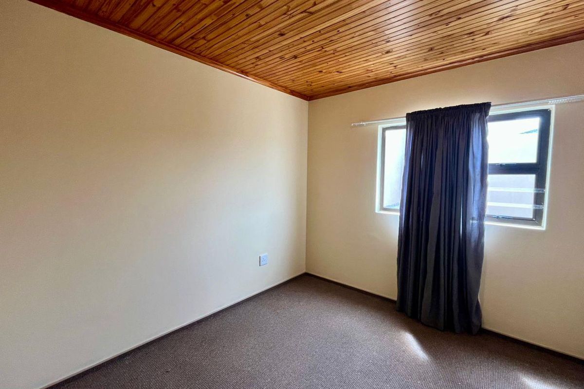 A simple bedroom featuring a timber-paneled ceiling, neutral walls, and carpeted flooring with a window providing natural light.