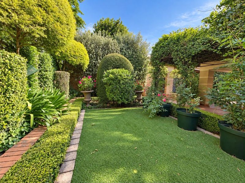 A well-maintained garden featuring an artificial lawn, neat brick edging, mature landscaped hedging, and a shaded pergola area.