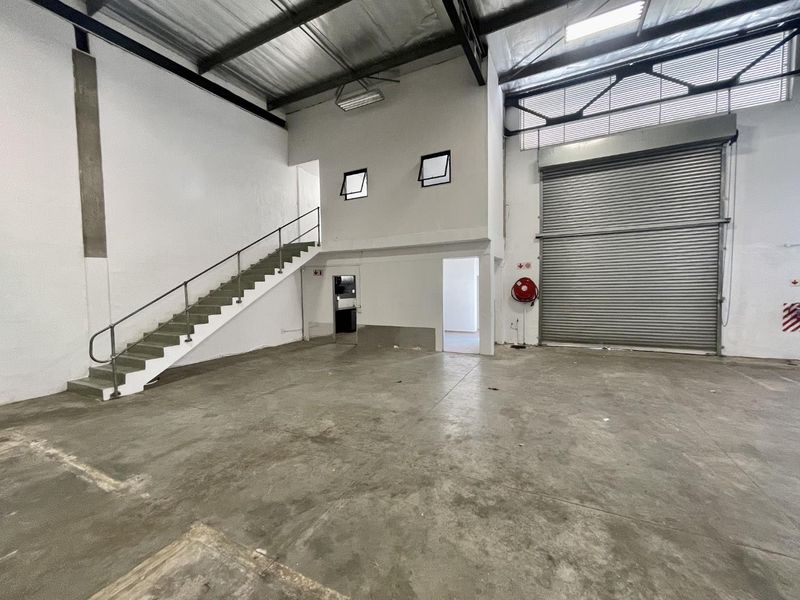 Spacious industrial warehouse unit featuring high ceilings with exposed trusses, a large roller shutter door, concrete flooring, and an internal staircase.