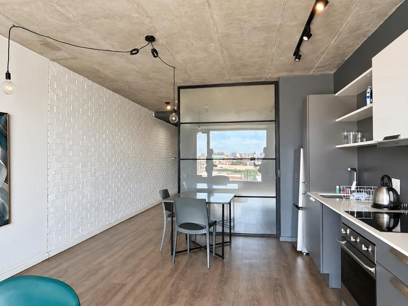 Modern open-plan apartment featuring an exposed concrete ceiling, white brick accent wall, sleek kitchen cabinetry, and a stylish black-framed glass partition.