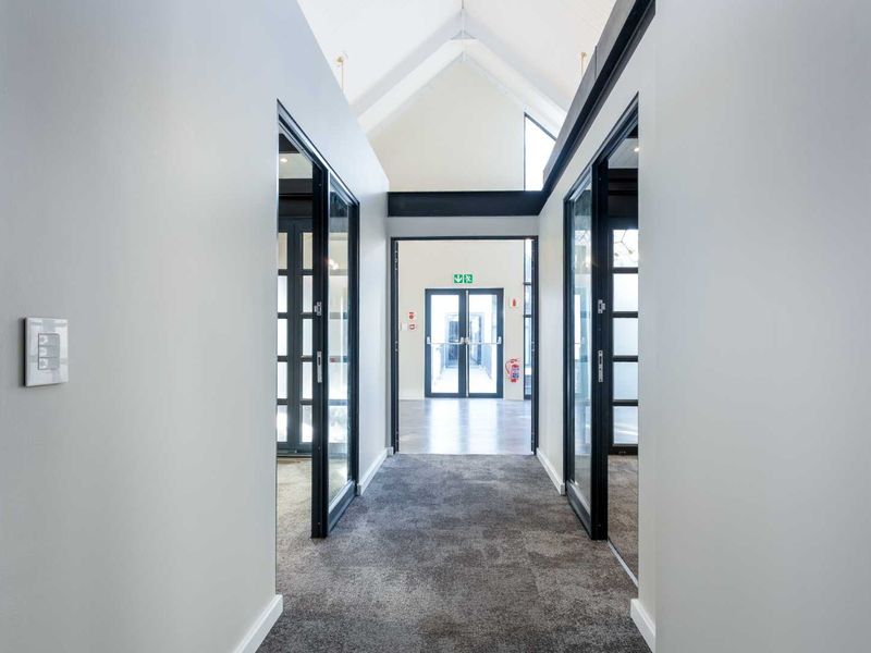 A bright, modern hallway featuring a high vaulted ceiling, carpeted flooring, and glass-paneled doors leading to adjacent office spaces.