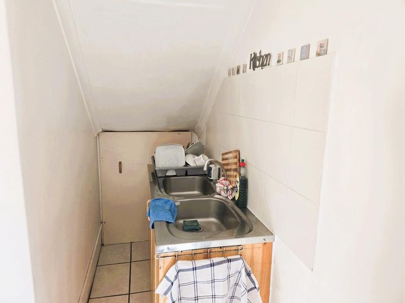 A compact scullery area featuring a stainless steel double sink, tiled splashback, and tiled flooring, situated under a sloped ceiling.