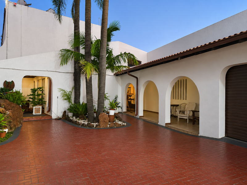 A well-maintained rear courtyard featuring a red brick paved area, arched covered patio spaces, and established palm trees.