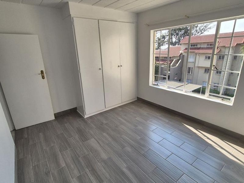 A bright, empty bedroom featuring modern wood-look tiled flooring, built-in cupboards, and a large window providing natural light and a view of the complex.