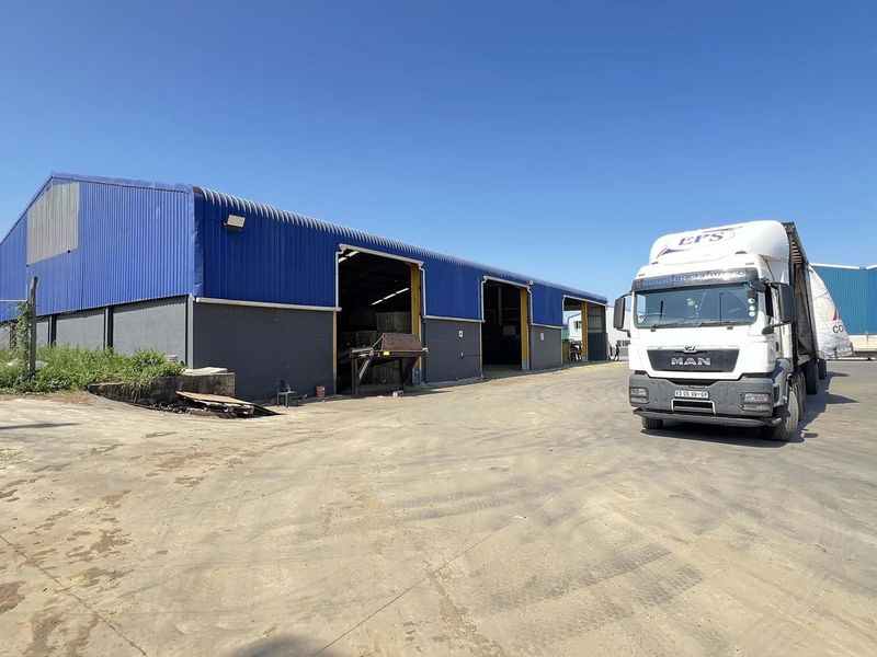 Industrial warehouse facility featuring large roller shutter access points, blue corrugated metal cladding, and a spacious concrete and asphalt yard area for logistics.