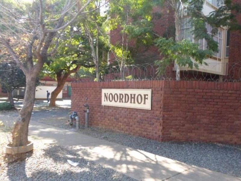 Street-level view of the Noordhof apartment complex entrance, featuring a brick boundary wall with security fencing and mature trees.