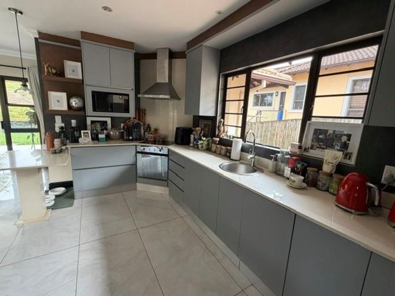 Modern kitchen featuring grey cabinetry, stone countertops, stainless steel appliances, and large windows providing natural light and garden views.