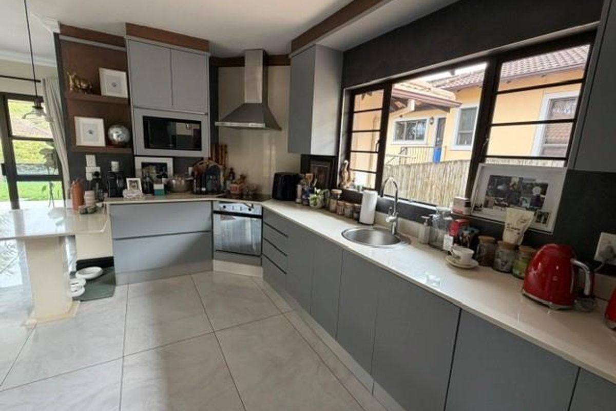 Modern kitchen featuring grey cabinetry, stone countertops, stainless steel appliances, and large windows providing natural light and garden views.