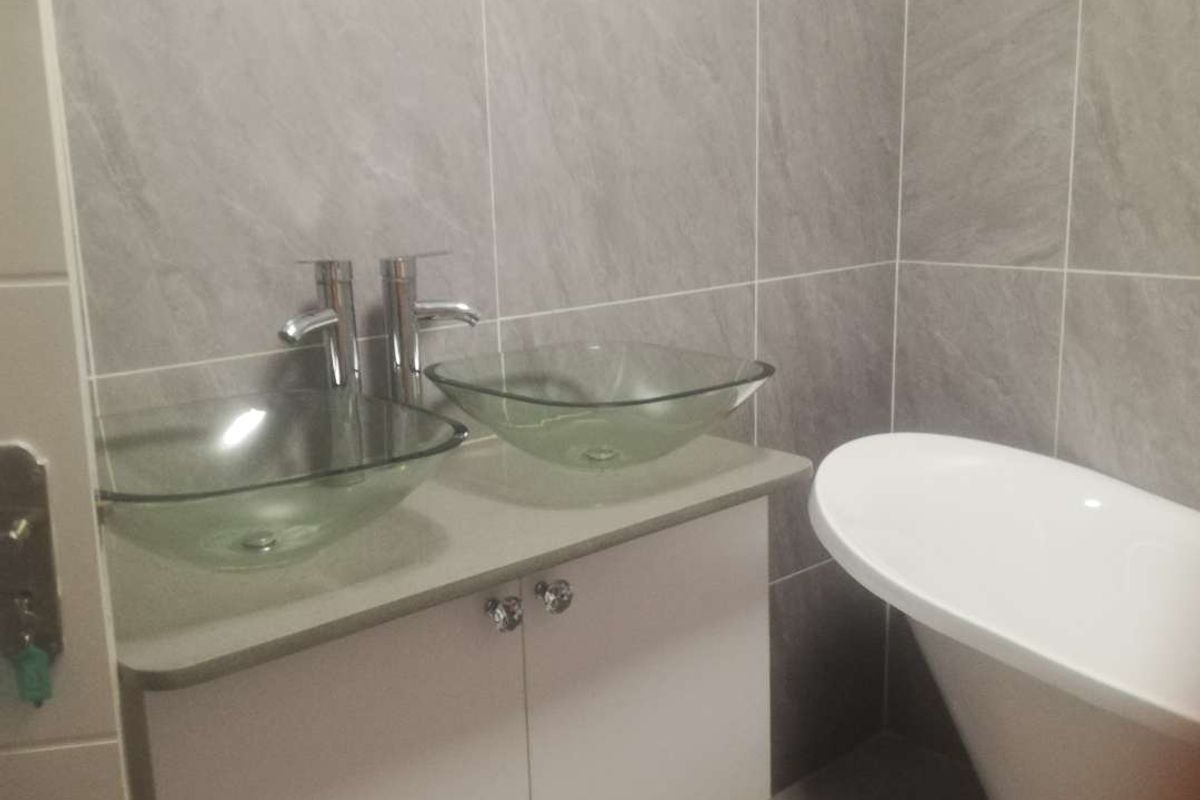 Modern bathroom featuring a double vanity with glass vessel sinks, chrome faucets, and a freestanding bathtub against grey tiled walls.