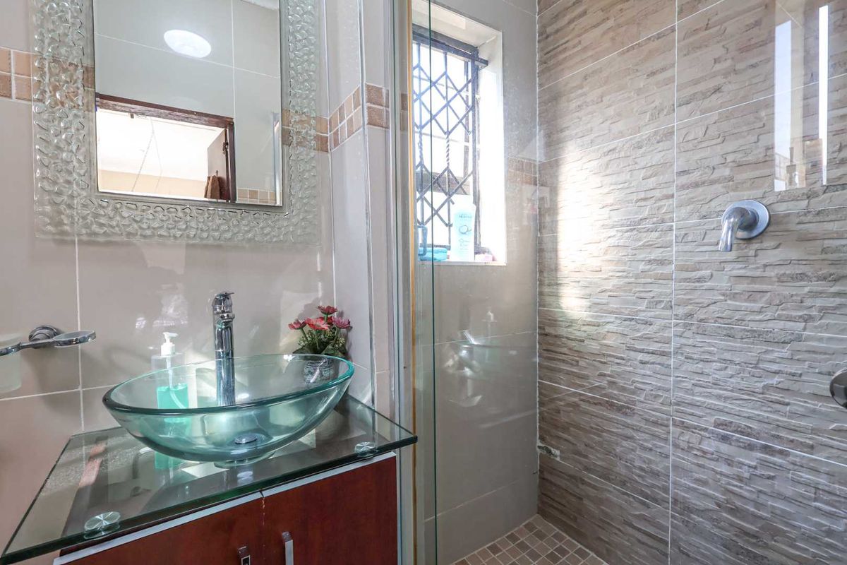 A modern bathroom featuring a glass vessel sink, vanity cabinet, wall-mounted mirror, and a walk-in shower with textured stone-look wall tiles.