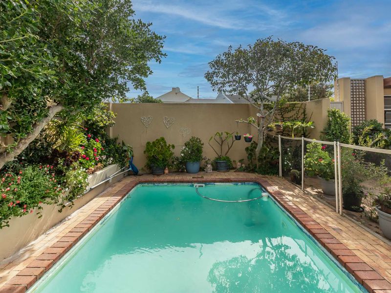 A private outdoor swimming pool area featuring a brick-edged pool, safety fencing, and established garden landscaping against a boundary wall.