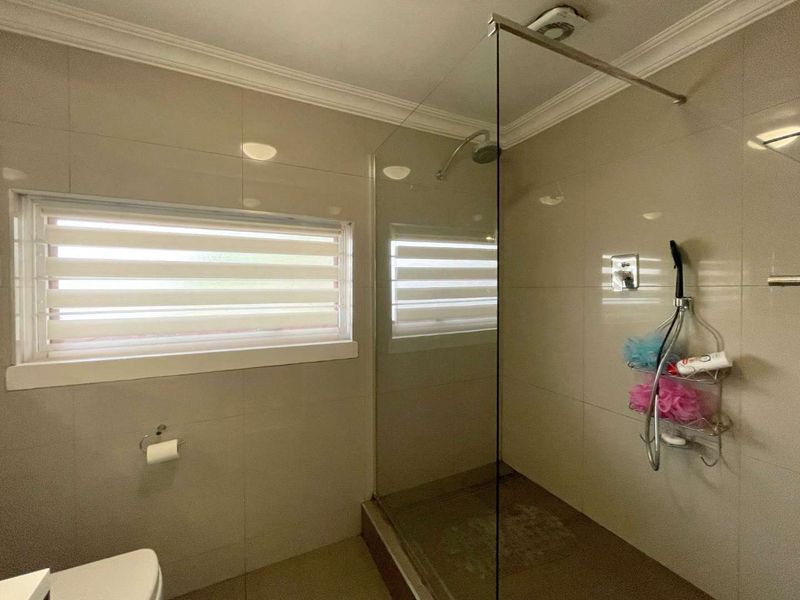 A modern bathroom featuring a glass-enclosed walk-in shower, neutral wall tiling, and a louver window for natural light and ventilation.
