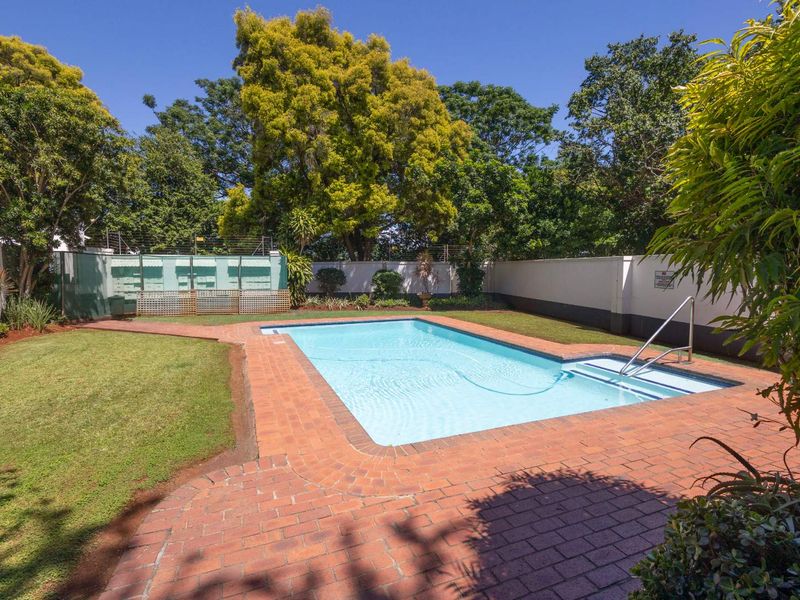 A well-maintained outdoor swimming pool area featuring brick paving, surrounding lawn, and secure perimeter walling with electric fencing.