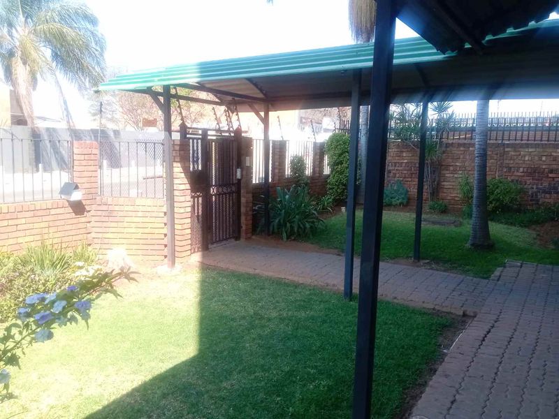A well-maintained front garden area featuring a paved walkway, a covered carport, and secure brick boundary walls with a metal gate.