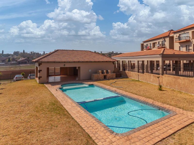 Outdoor pool area featuring a swimming pool, covered patio, and built-in braai facility within a fenced complex setting.
