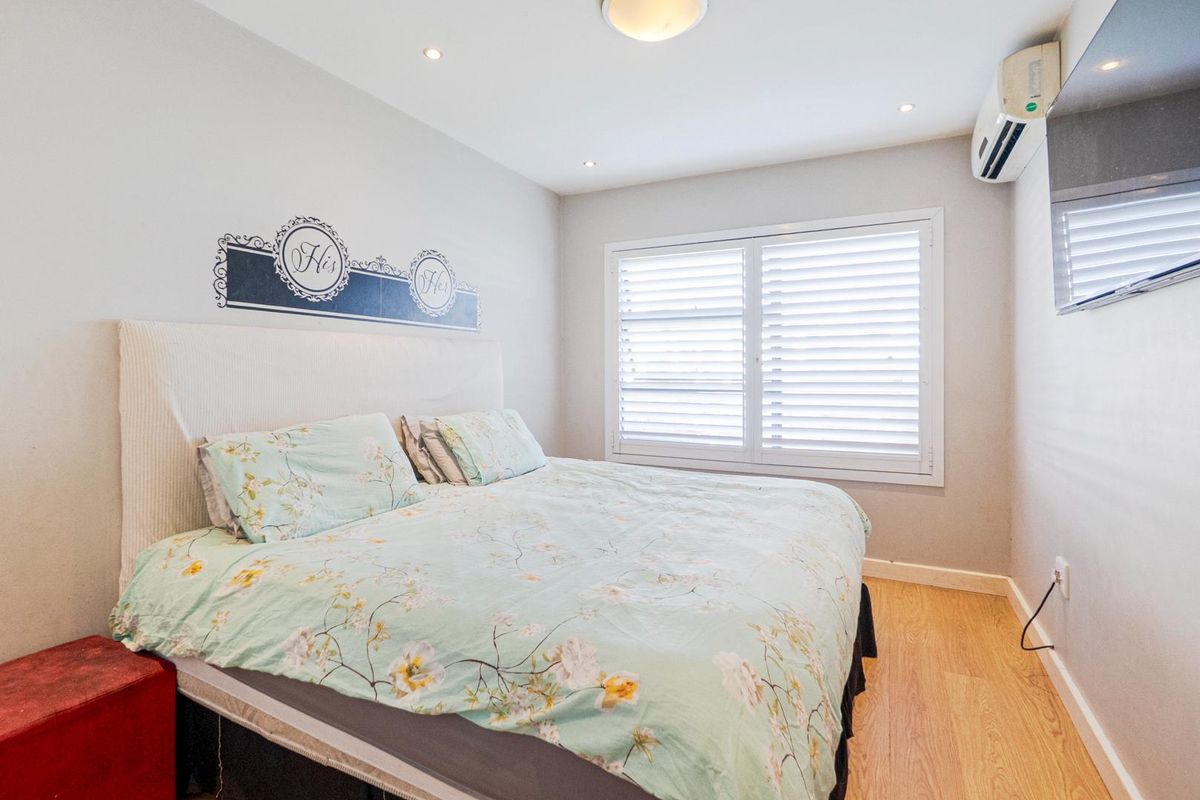 A well-maintained bedroom featuring light wood-look flooring, plantation shutters, recessed downlighting, and a wall-mounted air conditioning unit.