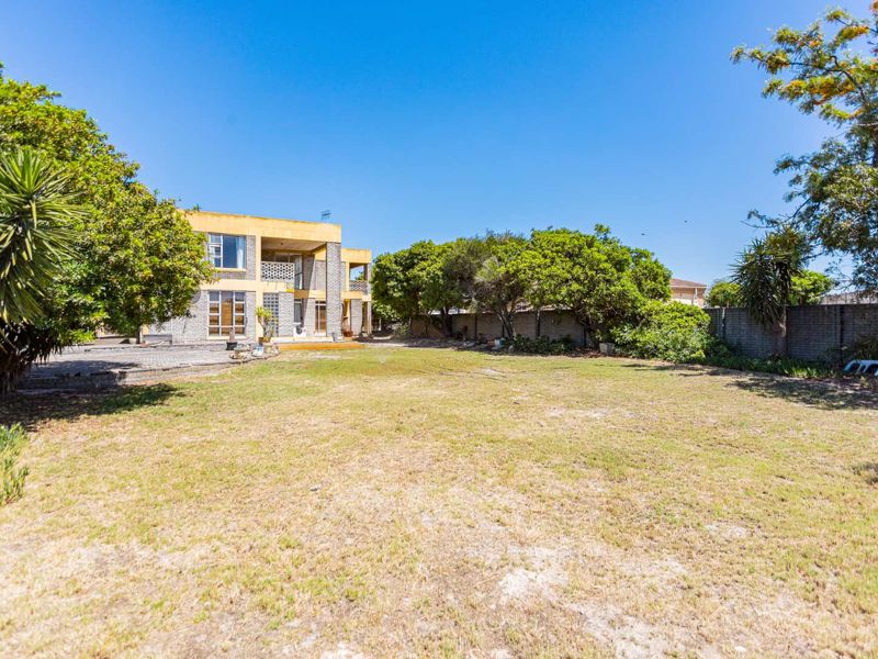 Spacious rear garden featuring a large lawn, mature trees, brick boundary walls, and a multi-level house facade with a paved patio area.
