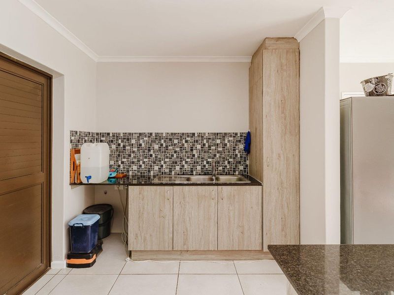 A functional scullery area featuring wood-look cabinetry, a stainless steel sink, granite-style countertops, and a decorative mosaic tile backsplash.