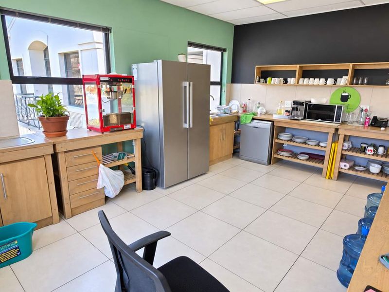A functional office kitchen area featuring tiled flooring, built-in wooden cabinetry, stainless steel sink, and open wall-mounted shelving for storage.