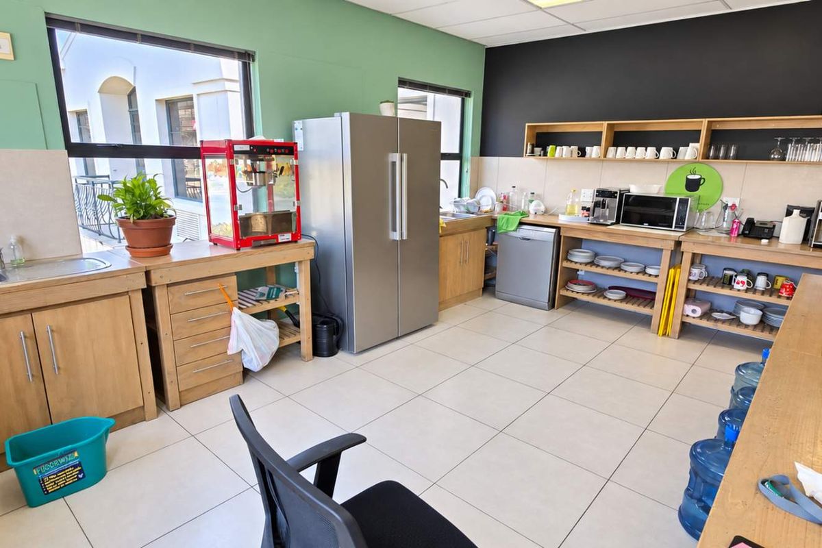 A functional office kitchen area featuring tiled flooring, built-in wooden cabinetry, stainless steel sink, and open wall-mounted shelving for storage.