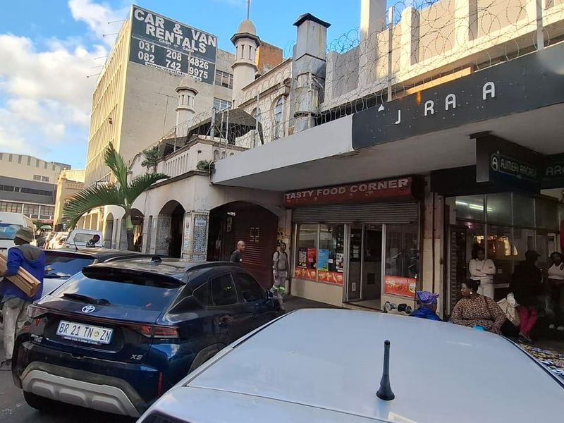 Street-level view of a commercial building facade featuring a food corner storefront and arcade entrance in a busy urban environment.
