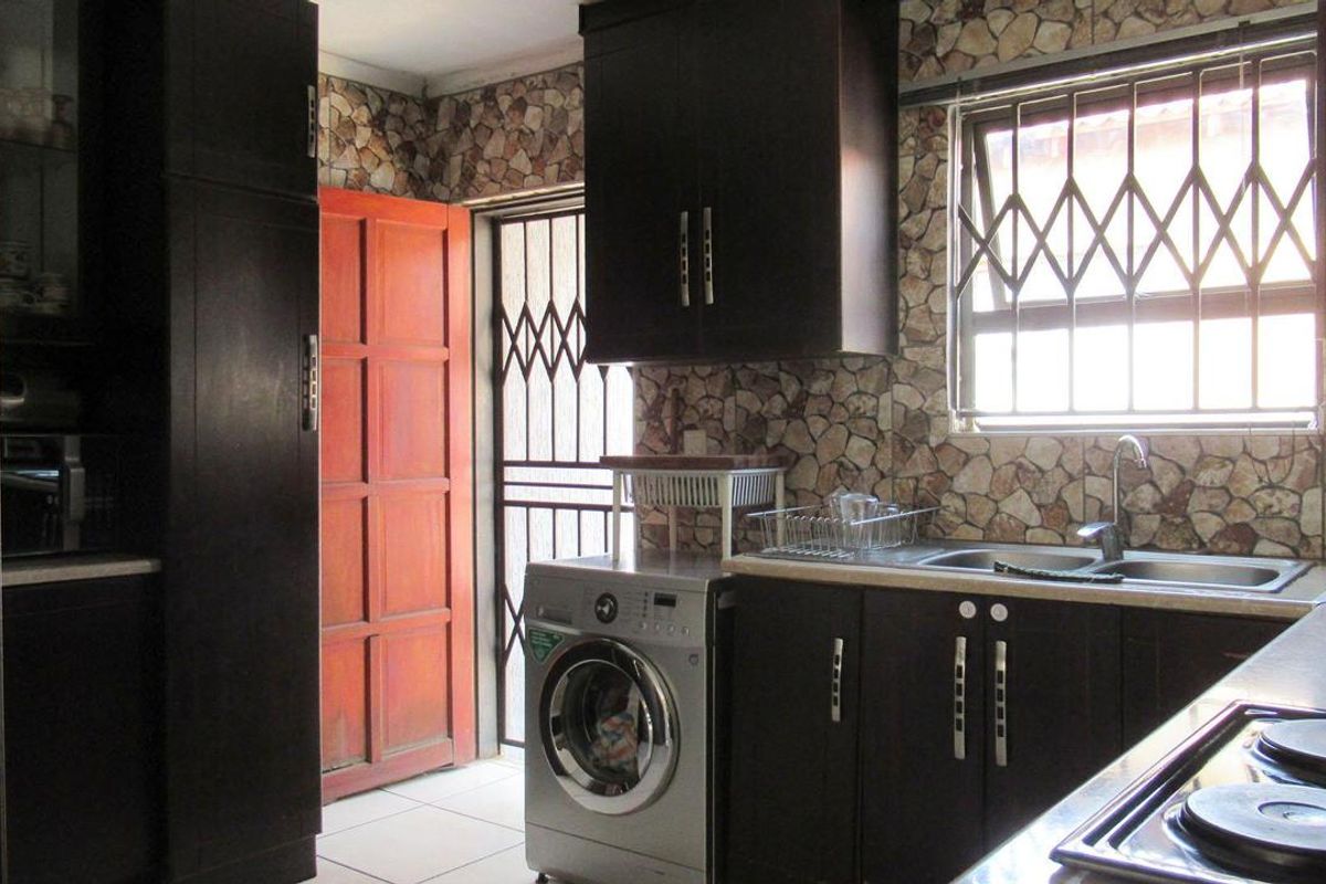 A functional kitchen featuring dark cabinetry, stone-effect wall cladding, a stainless steel sink, and a built-in hob, with direct access to an exterior door.