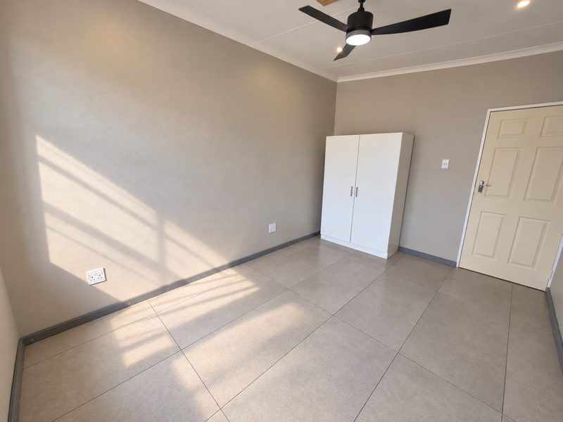 A bright, empty bedroom featuring neutral wall tones, large floor tiles, a modern ceiling fan, and a built-in white cupboard.