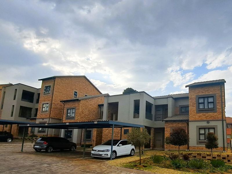 Exterior view of a multi-story apartment complex featuring brick and plaster facade, covered parking carports, and a paved driveway with landscaped garden areas.