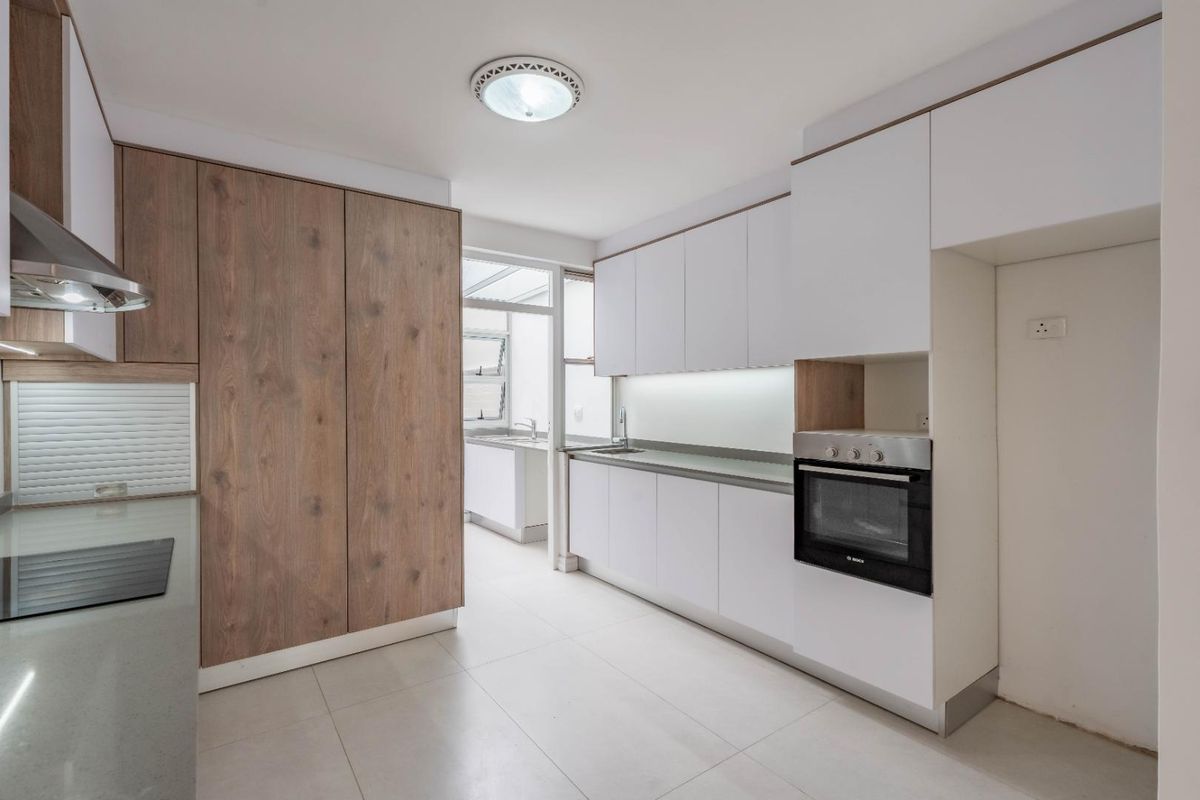 Modern kitchen featuring sleek white cabinetry, wood-grain pantry storage, integrated oven, extractor fan, and stone countertops, leading into a bright scullery area.