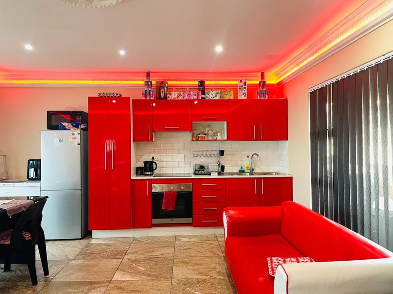 Open-plan living area featuring a modern kitchen with high-gloss red cabinetry, built-in appliances, subway tile backsplash, and ambient cove lighting.