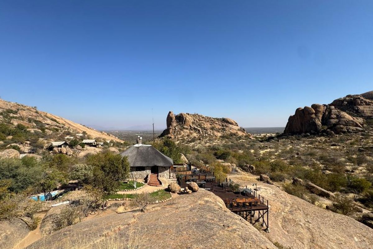 An elevated aerial view of a lodge featuring a traditional thatched roof structure, stone exterior, and a raised timber deck overlooking a rocky landscape.