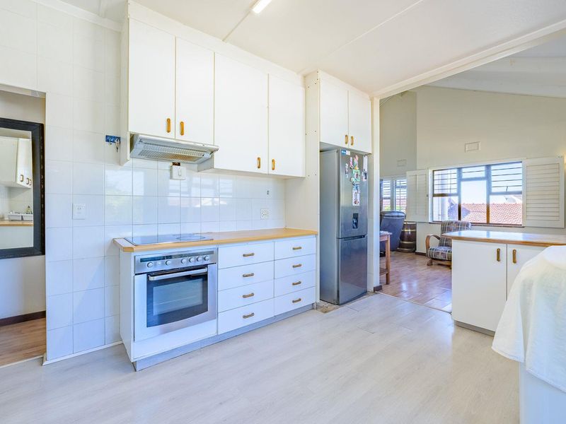 A functional kitchen featuring white cabinetry, a built-in oven and hob, tiled splashbacks, and light-toned laminate flooring, flowing into an adjacent living area.