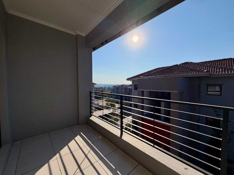 A spacious covered balcony featuring tiled flooring and a modern metal balustrade, offering an open view of the surrounding residential complex.