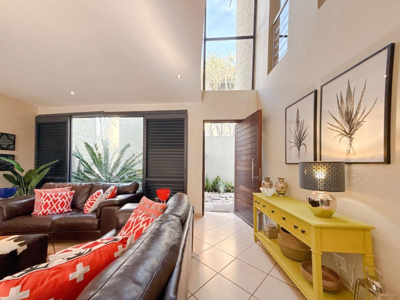 Spacious living area featuring double volume ceilings, large windows with sliding shutters, and direct access to an outdoor courtyard through a timber entrance door.