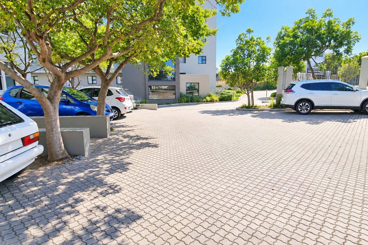 A well-maintained paved parking area and driveway leading to the office building entrance, featuring mature trees and concrete landscaping elements.